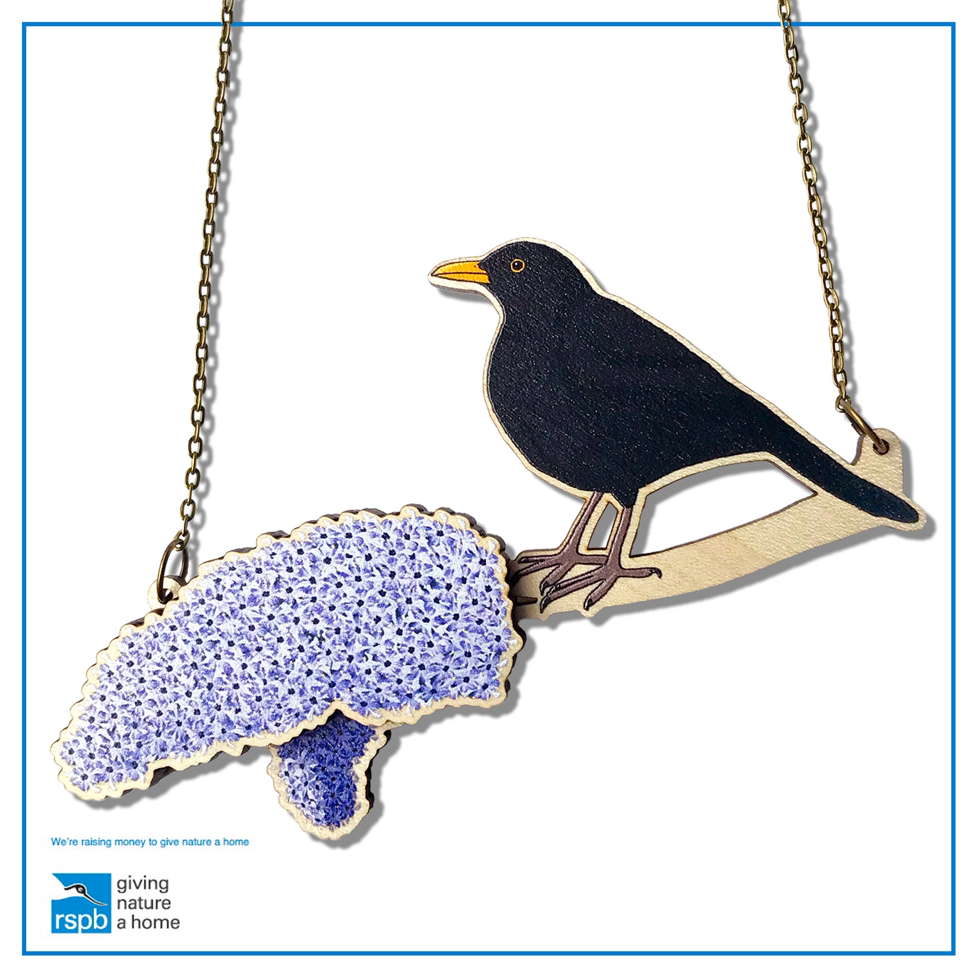 A laser cut wooden charm necklace showing an illustration of a male blackbird perching on a branch with lilac blossoms by loadofolbobbins. The lilac blossom flowers stand proud of the necklace in a 3D relief effect.  The wooden necklace is hanging from an antique bronze cable chain.
