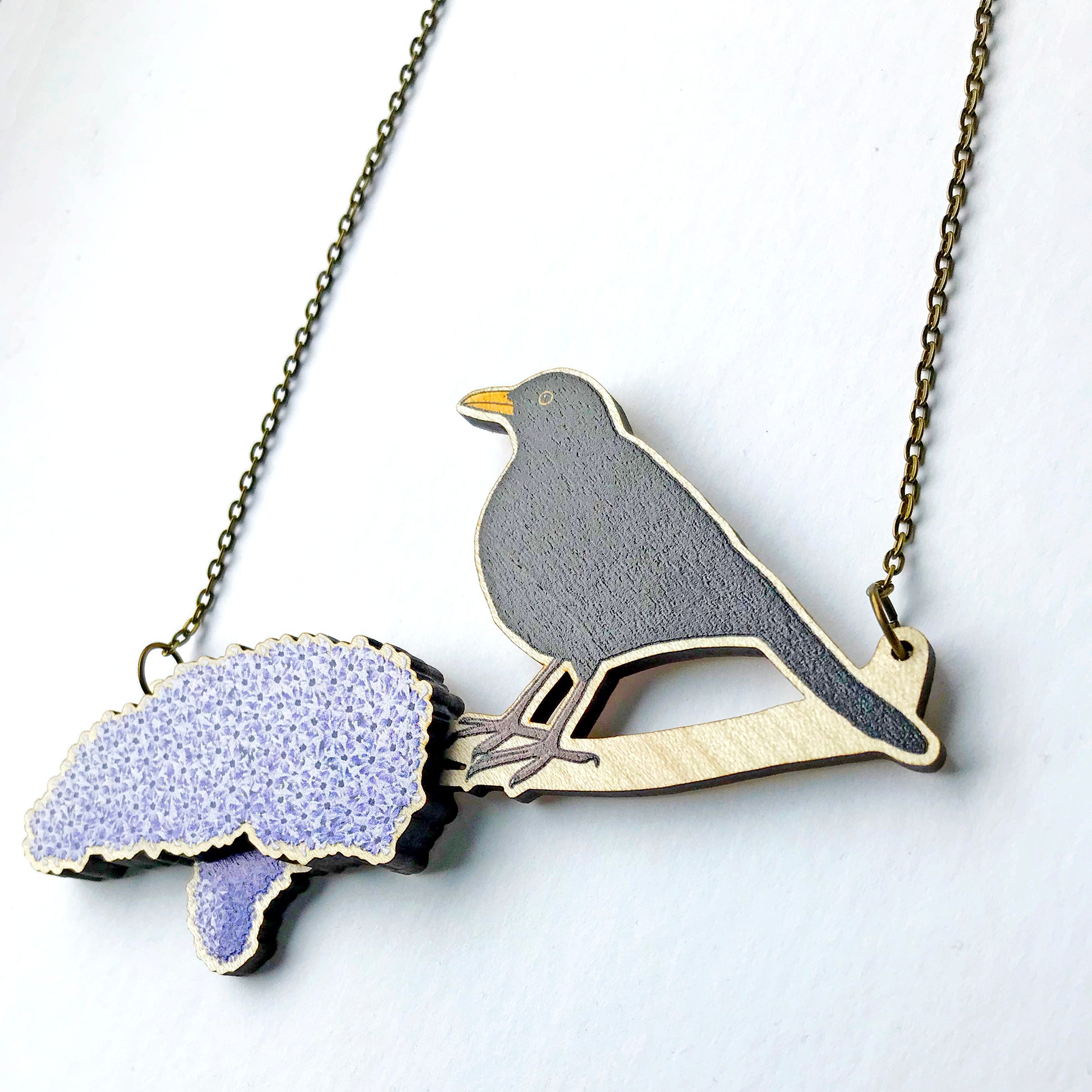 A laser cut wooden charm necklace showing an illustration of a male blackbird perching on a branch with lilac blossoms by loadofolbobbins. The lilac blossom flowers stand proud of the necklace in a 3D relief effect.  The wooden necklace is hanging from an antique bronze cable chain.