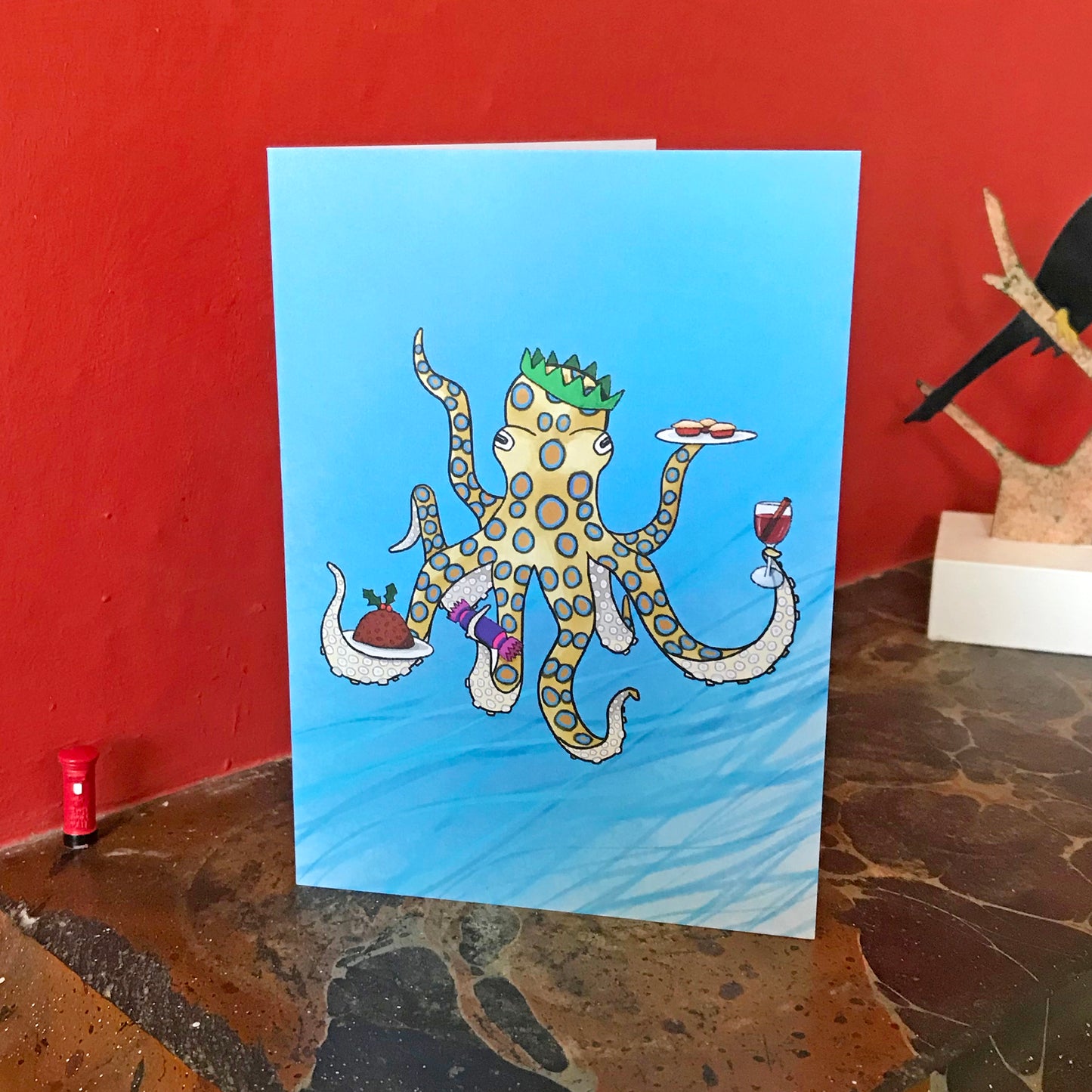 A greetings card by Loadofolbobbins. It shows a digital illustration of a Blue-ringed octopus underwater and clearly having managed to find the Christmas buffet. With a green paper crown on it's head, in four of it's tentacles it's holding festive items including a Christmas pudding, a Christmas crack, some mulled wine and a plate of mince pies. The card is on a mantelpiece.