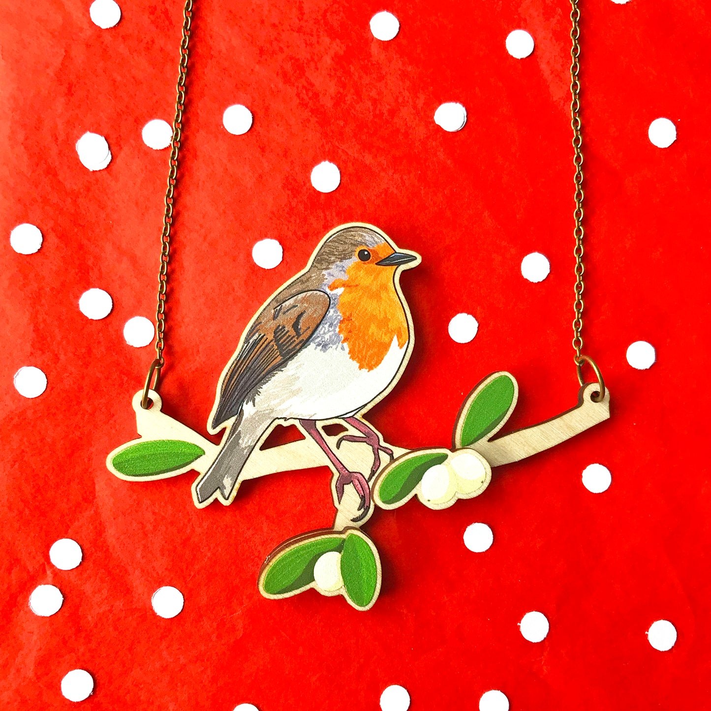 A laser cut wooden charm necklace showing an illustration of a robin bird perching on a branch with mistletoe by loadofolbobbins. The mistletoe berries and leaves stand proud of the necklace in a 3D relief effect.  The wooden necklace is hanging from an antique bronze cable chain and displayed against a red snowy background.