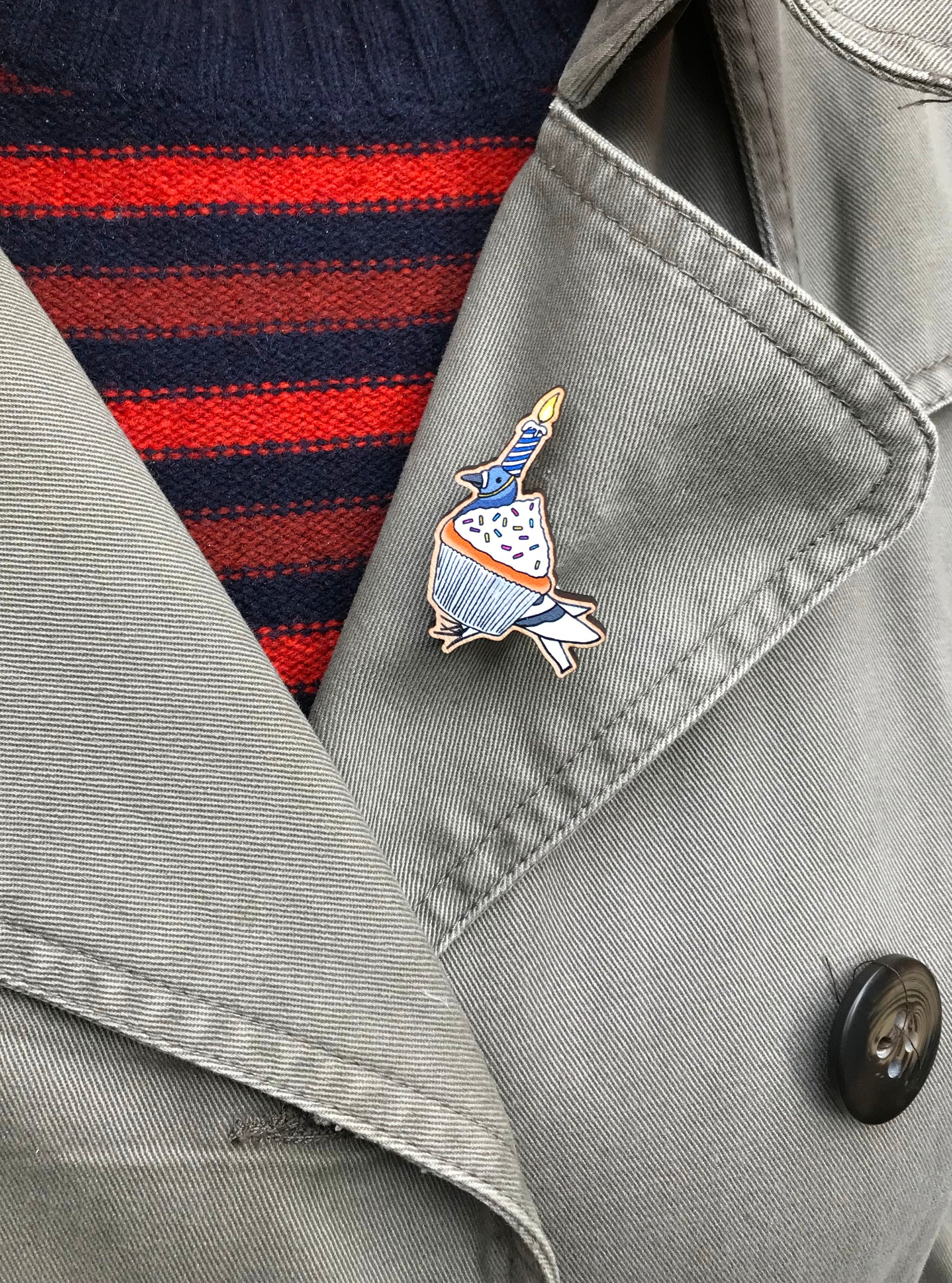 A laser cut wooden lapel pin showing an original illustration by Loadofolbobbins of a pigeon dressed up as a cupcake with a lit birthday candle strapped to it's head. The style of the illustration is simplified with flat colours and a bold black outline. The pin is mounted on a blue backing card that says Coosplay with Fred the pigeon. The pin is shown on the lapel of a jacket.