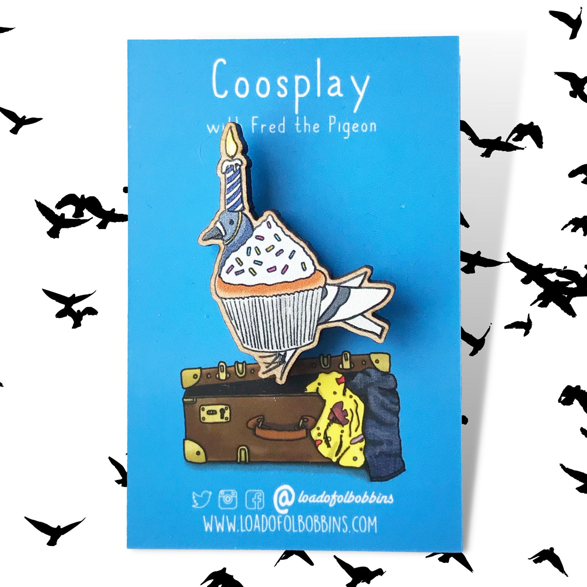 A laser cut wooden lapel pin showing an original illustration by Loadofolbobbins of a pigeon dressed up as a cupcake with a lit birthday candle strapped to it's head. The style of the illustration is simplified with flat colours and a bold black outline. The pin is mounted on a blue backing card that says Coosplay with Fred the pigeon.