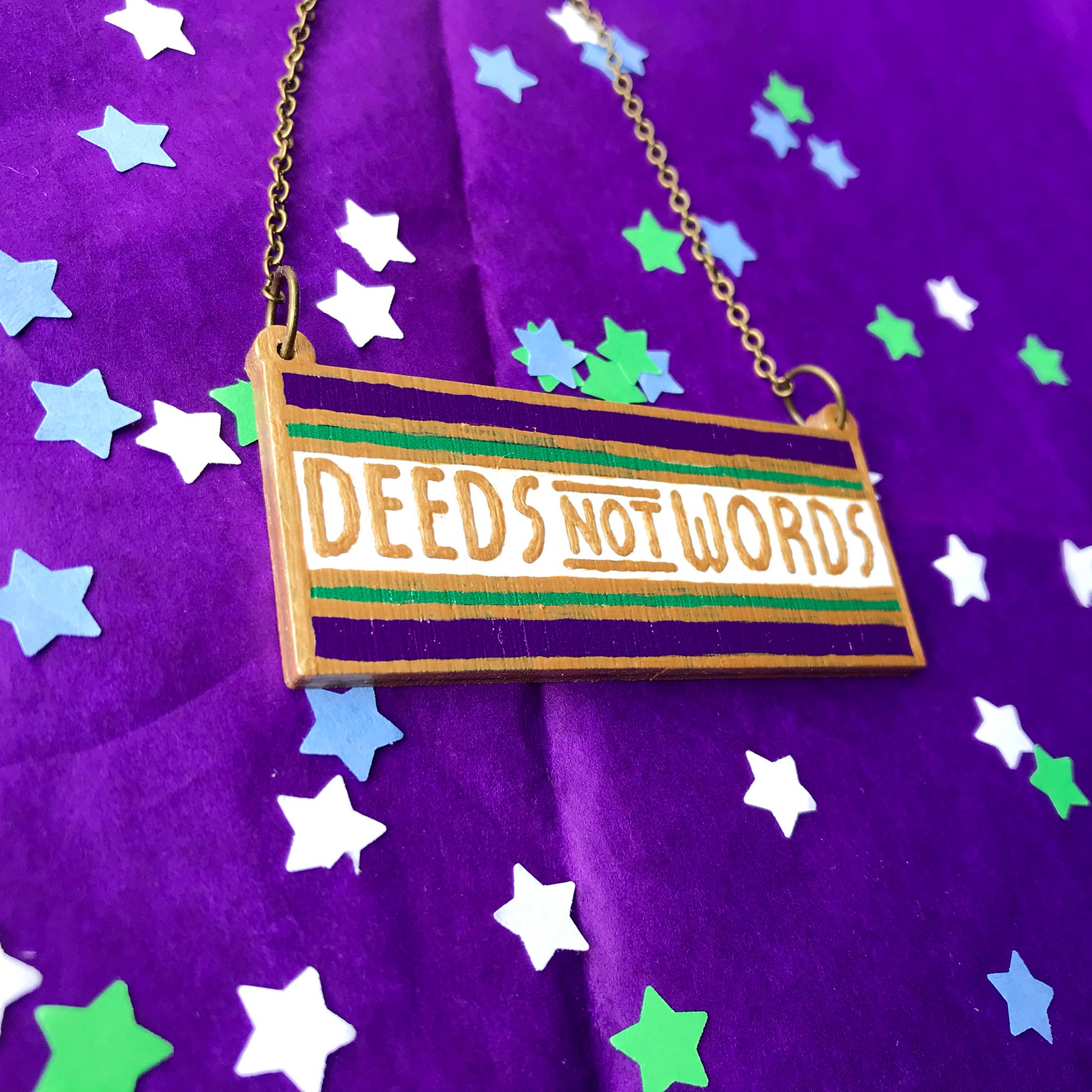 An oblong necklace charm with deeds not words written against a white stripe and sandwiched between a thin green stripe and thicker purple stripe. The line borders and text have been hand painted in gold paint. It is hung from an antique bronze cable chain.
