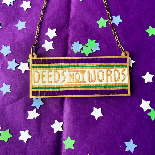 An oblong necklace charm with deeds not words written against a white stripe and sandwiched between a thin green stripe and thicker purple stripe. The line borders and text have been hand painted in gold paint. It is hung from an antique bronze cable chain.