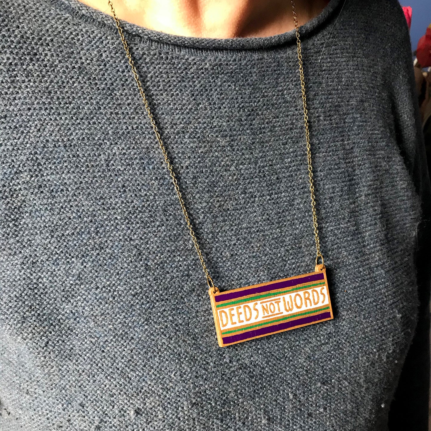 An oblong necklace charm with deeds not words written against a white stripe and sandwiched between a thin green stripe and thicker purple stripe. The line borders and text have been hand painted in gold paint. It is hung from an antique bronze cable chain. It's worn against a blue jumper.