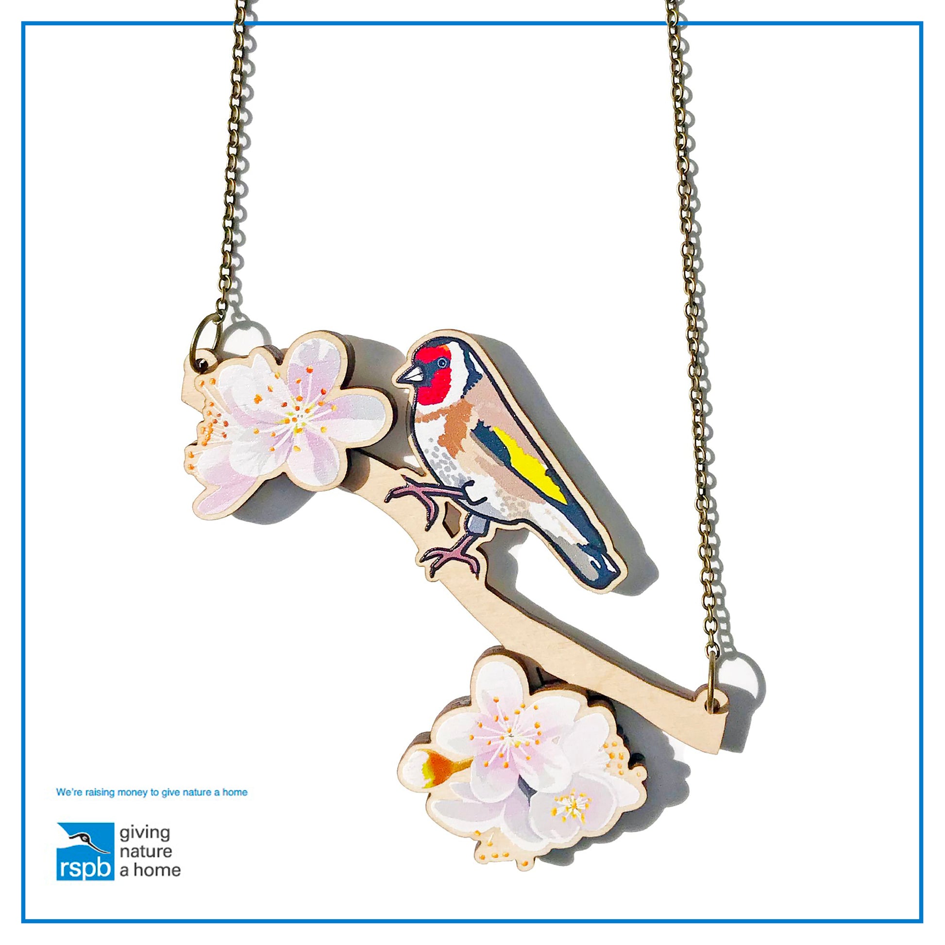 A laser cut wooden charm necklace showing an illustration of a goldfinch bird perching on a branch with cherry blossoms by loadofolbobbins. The cherry blossom flowers stand proud of the necklace in a 3D relief effect.  The wooden necklace is hanging from an antique bronze cable chain.