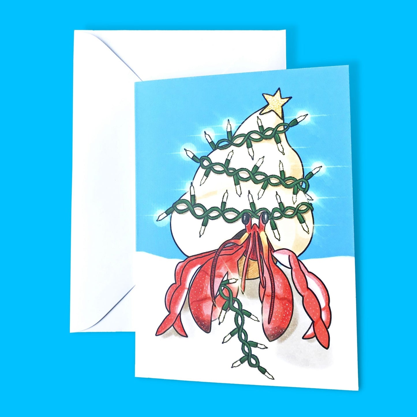An A6 greetings card showing an illustration by Loadofolbobbins of a hermit crab standing on a sandy beach under vibrant blue sky. Wrapped round their shell is a string of glowing fairy lights that they hold in their right claw. On top of their shell is a glittery gold star, like a Christmas tree. 