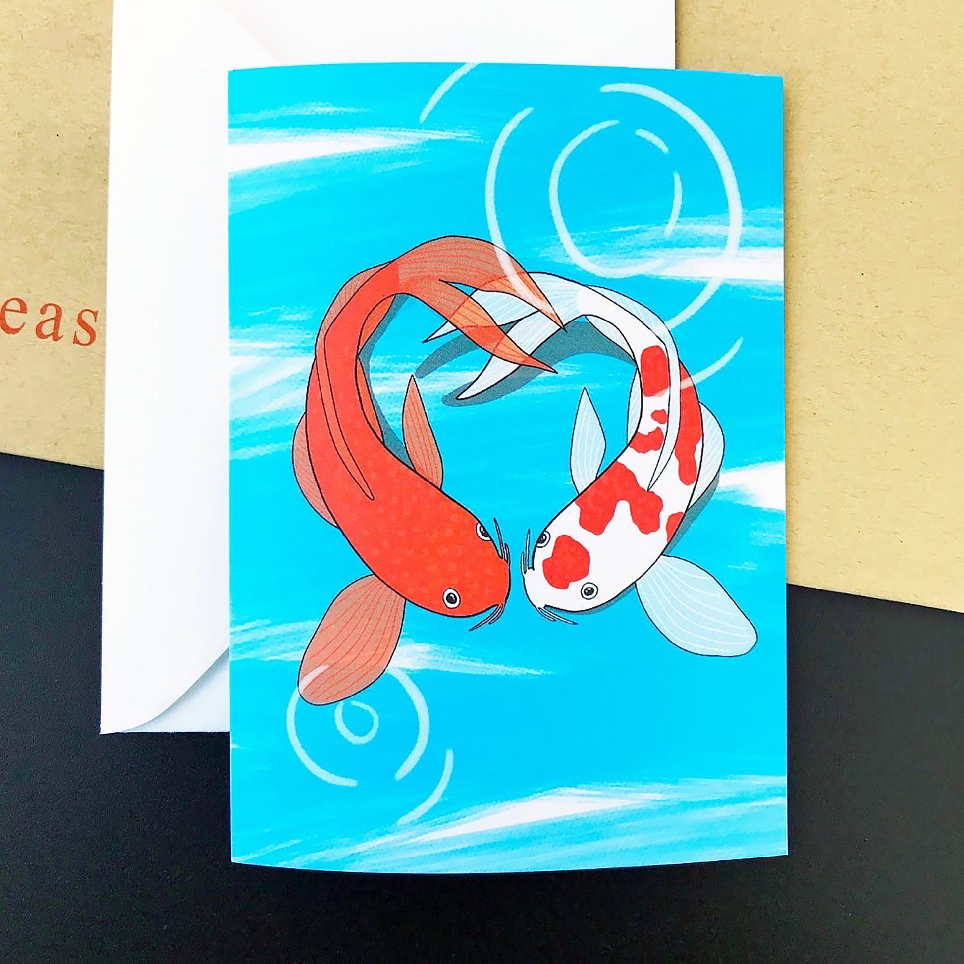 An A6 greetings card with an illustration by Loadofolbobbins of two koi fish facing each other in a pond and forming the shape of a heart. The koi on the left is a vibrant orange and the koi on the right is white with vibrant orange splodges. The background is an energetic light blue line zig zagging down the image with some white showing through. Light white circular marks show ripples on the surface of the water.