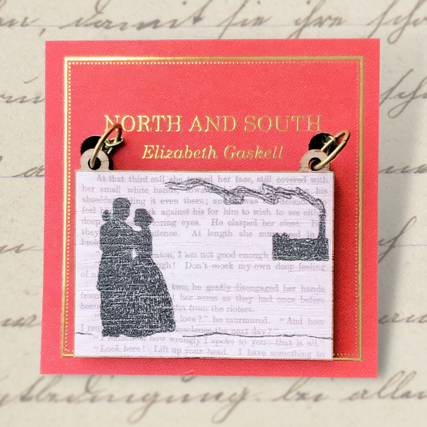 A laser cut wooden necklace showing a silhouette scene a couple in Victorian dress embracing and a factory in the background with a billowing chimney. It is inspired by the novel North and South by Elizabeth Gaskell and is printed with the text from the novel behind it. The necklace is mounted on a red backing card with gold foil lettering detail and made to look like the front of a book.