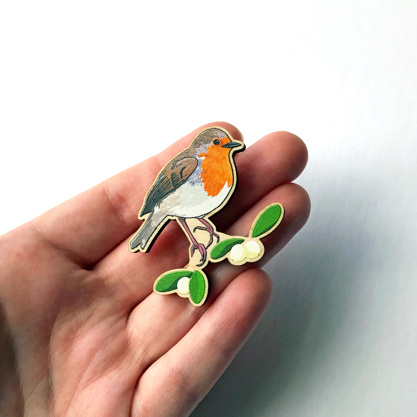 A laser cut wooden brooch showing an illustration by Loadofolbobbins of a robin bird perched on some mistletoe. The brooch is held in a hand for scale.