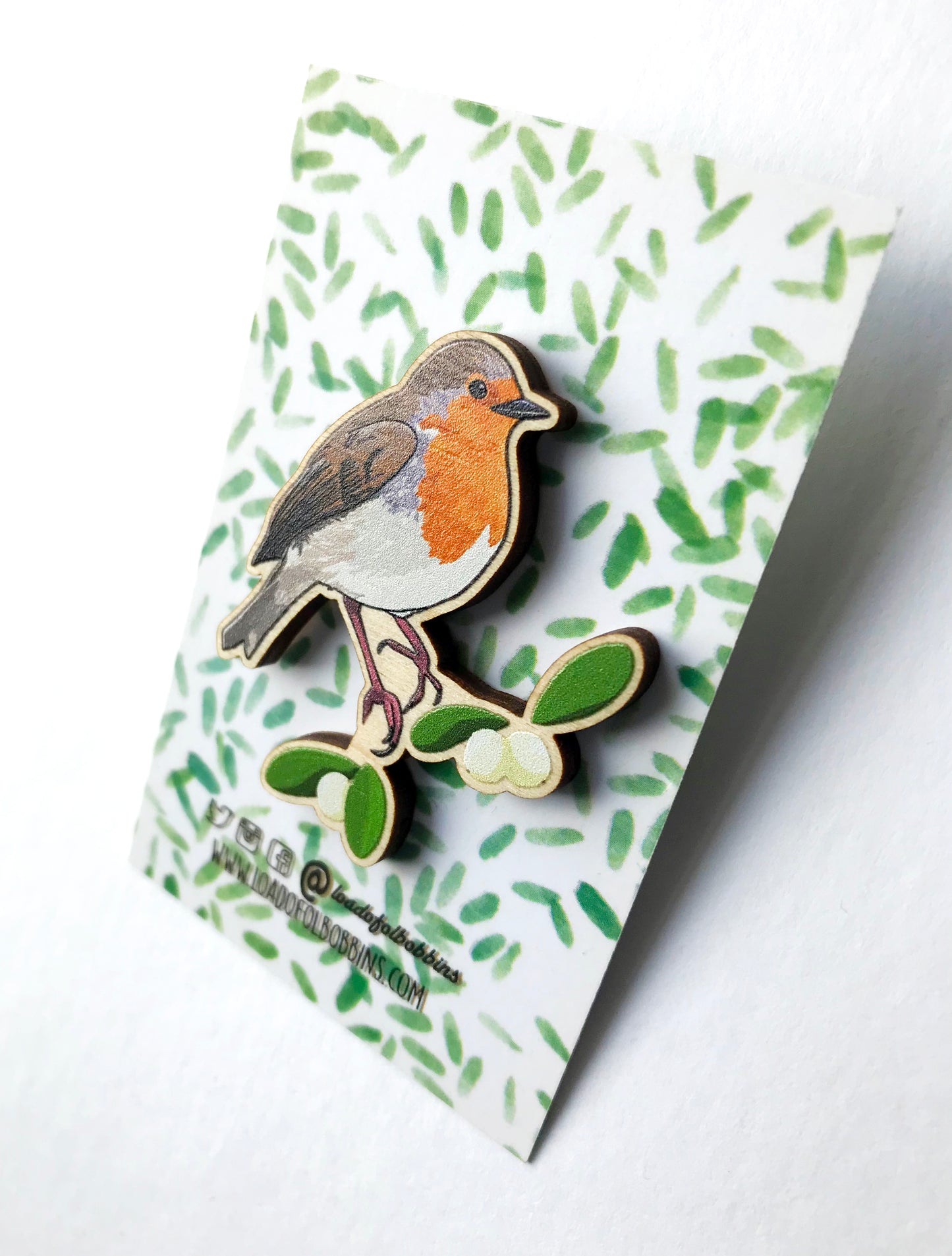 A laser cut wooden brooch showing an illustration by Loadofolbobbins of a robin bird perched on some mistletoe. The brooch is mounted on a backing card with a repeat abstract watercolour leaf pattern.