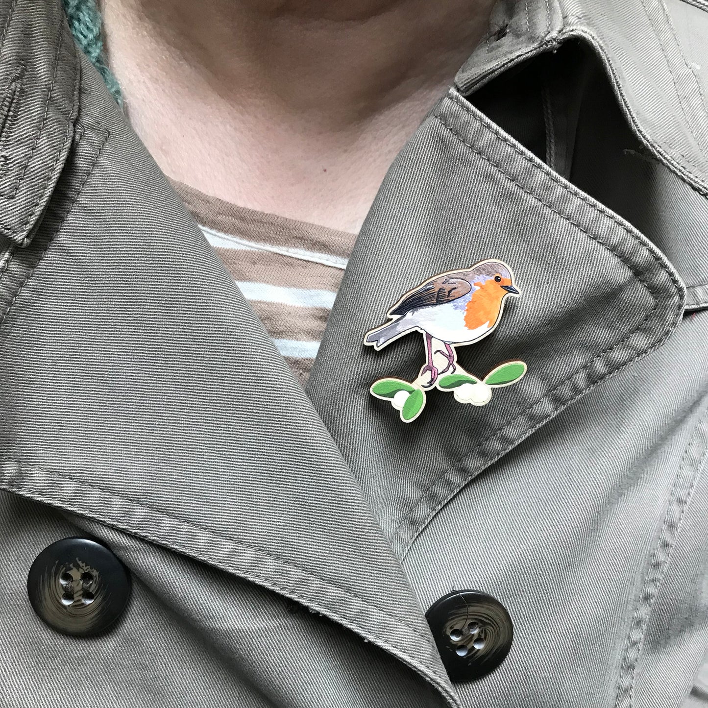 A laser cut wooden brooch showing an illustration by Loadofolbobbins of a robin bird perched on some mistletoe. The brooch is mounted on a backing card with a repeat abstract watercolour leaf pattern. It is shown on the lapel of a jecket.