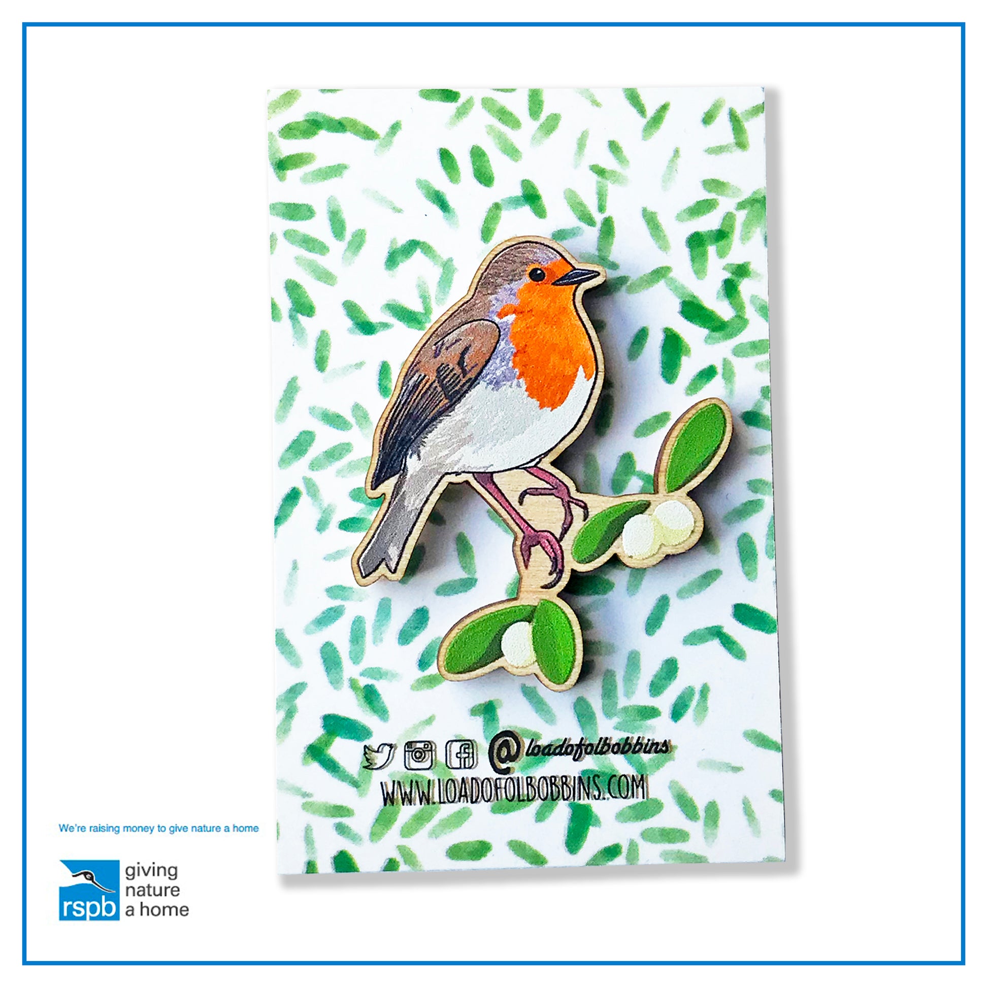 A laser cut wooden brooch showing an illustration by Loadofolbobbins of a robin bird perched on some mistletoe. The brooch is mounted on a backing card with a repeat abstract watercolour leaf pattern.