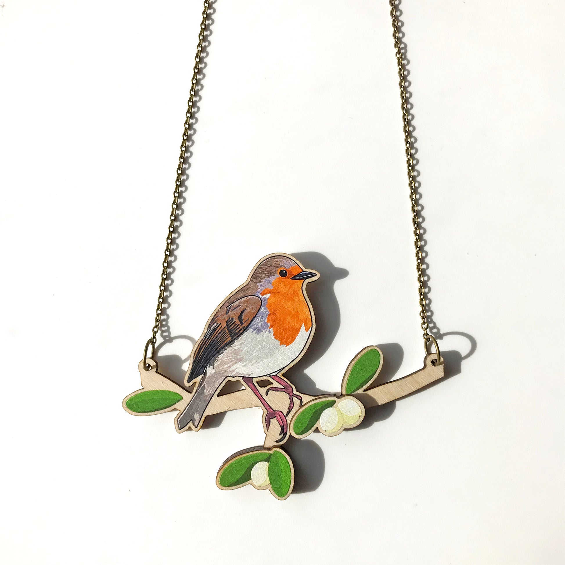 A laser cut wooden charm necklace showing an illustration of a robin bird perching on a branch with mistletoe by loadofolbobbins. The mistletoe berries and leaves stand proud of the necklace in a 3D relief effect.  The wooden necklace is hanging from an antique bronze cable chain.
