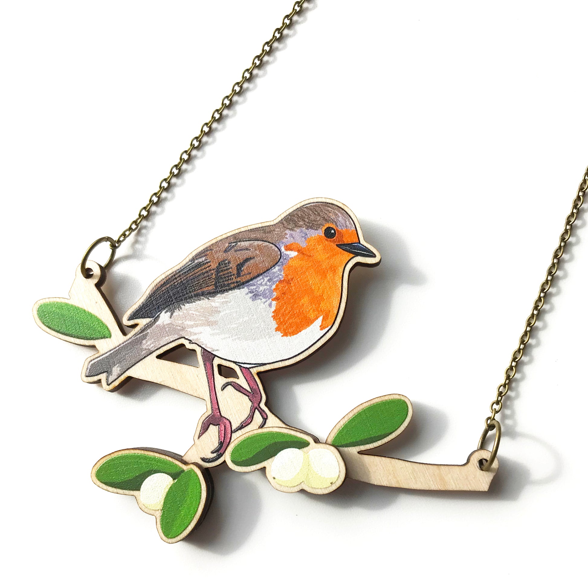 A laser cut wooden charm necklace showing an illustration of a robin bird perching on a branch with mistletoe by loadofolbobbins. The mistletoe berries and leaves stand proud of the necklace in a 3D relief effect.  The wooden necklace is hanging from an antique bronze cable chain.