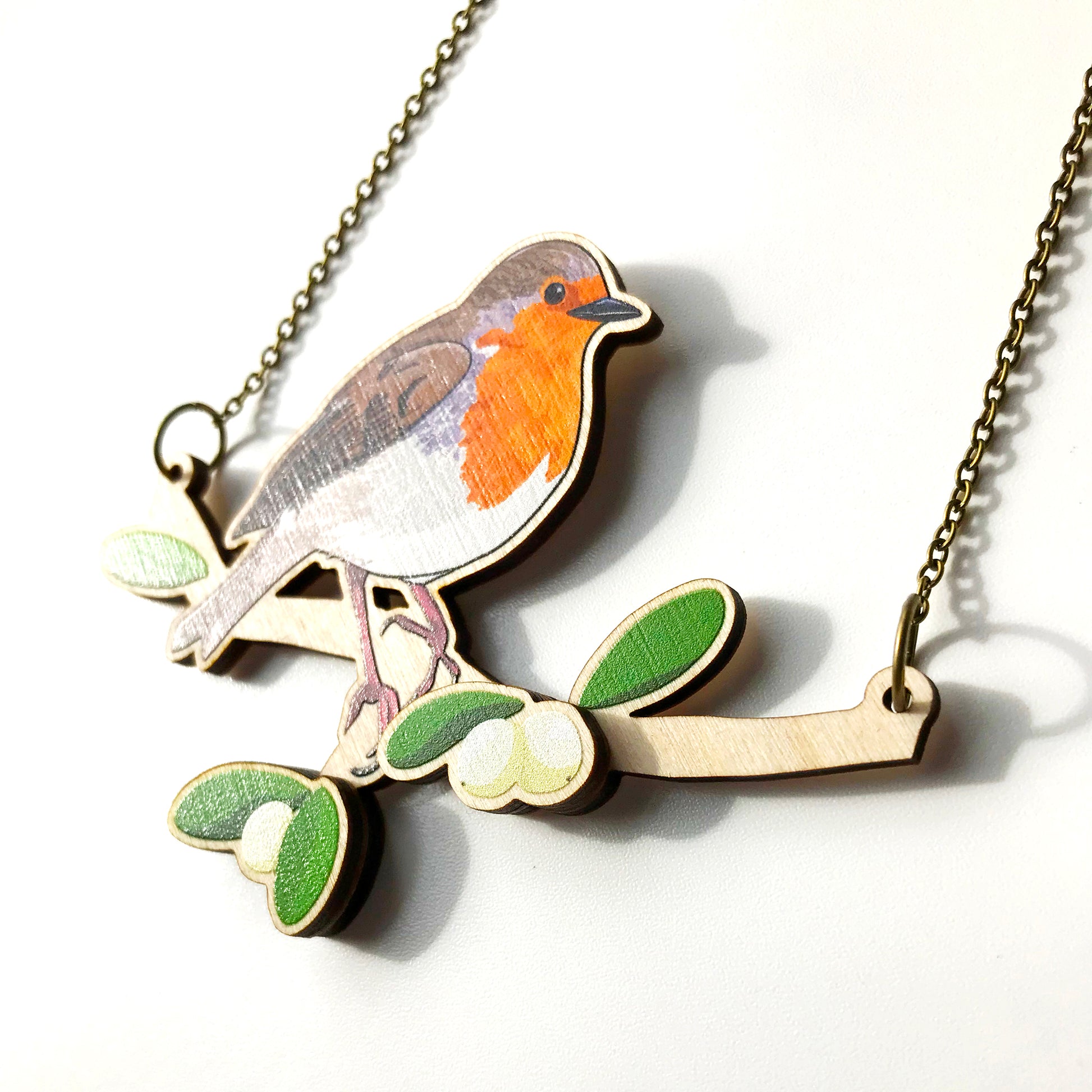 A laser cut wooden charm necklace showing an illustration of a robin bird perching on a branch with mistletoe by loadofolbobbins. The mistletoe berries and leaves stand proud of the necklace in a 3D relief effect.  The wooden necklace is hanging from an antique bronze cable chain.