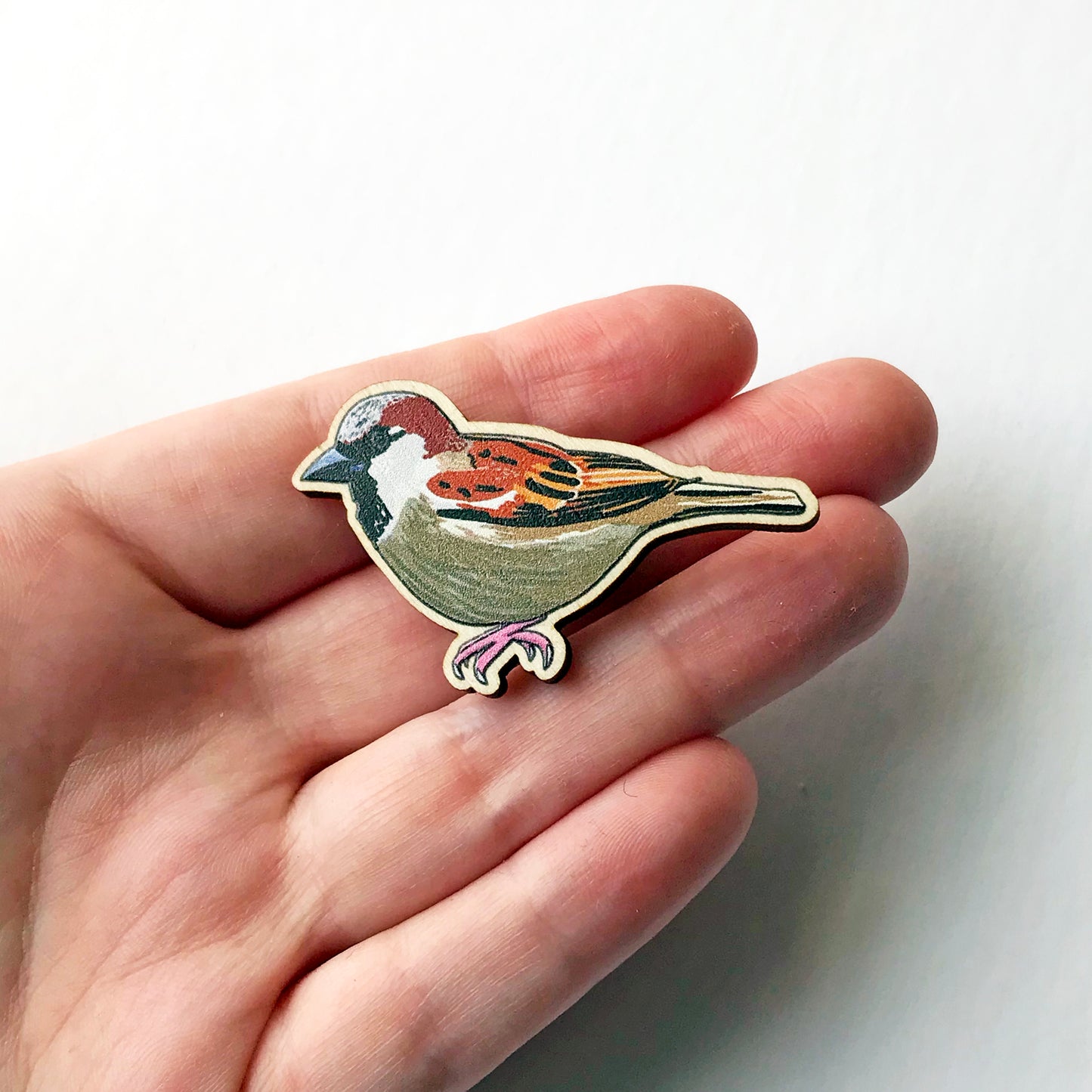 A laser cut wooden brooch showing an illustration by Loadofolbobbins of a male sparrow. The brooch is held in a hand for scale.