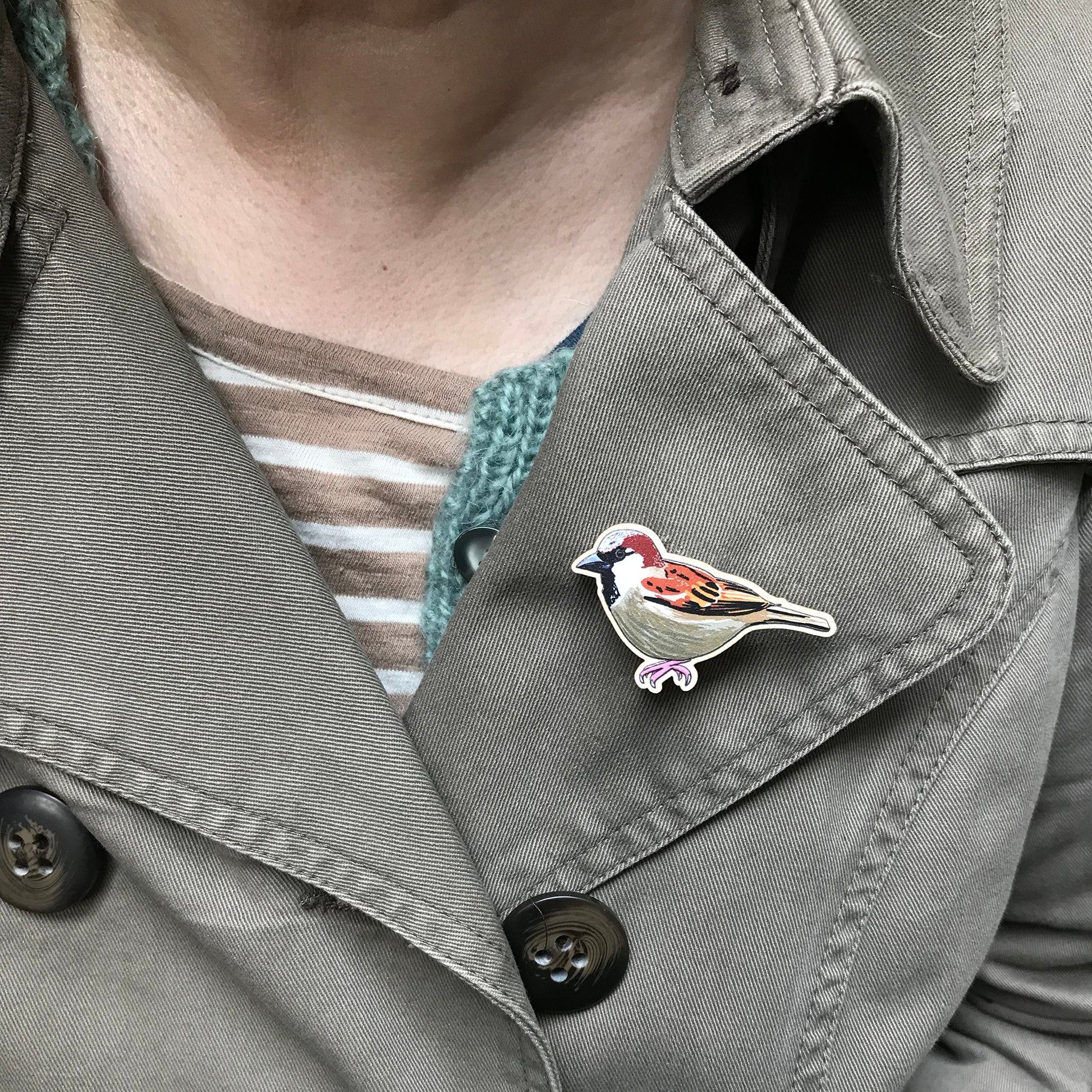 A laser cut wooden brooch showing an illustration by Loadofolbobbins of a male sparrow. The brooch is mounted on a backing card with a repeat abstract watercolour leaf pattern. It is shown on the lapel of a jacket.