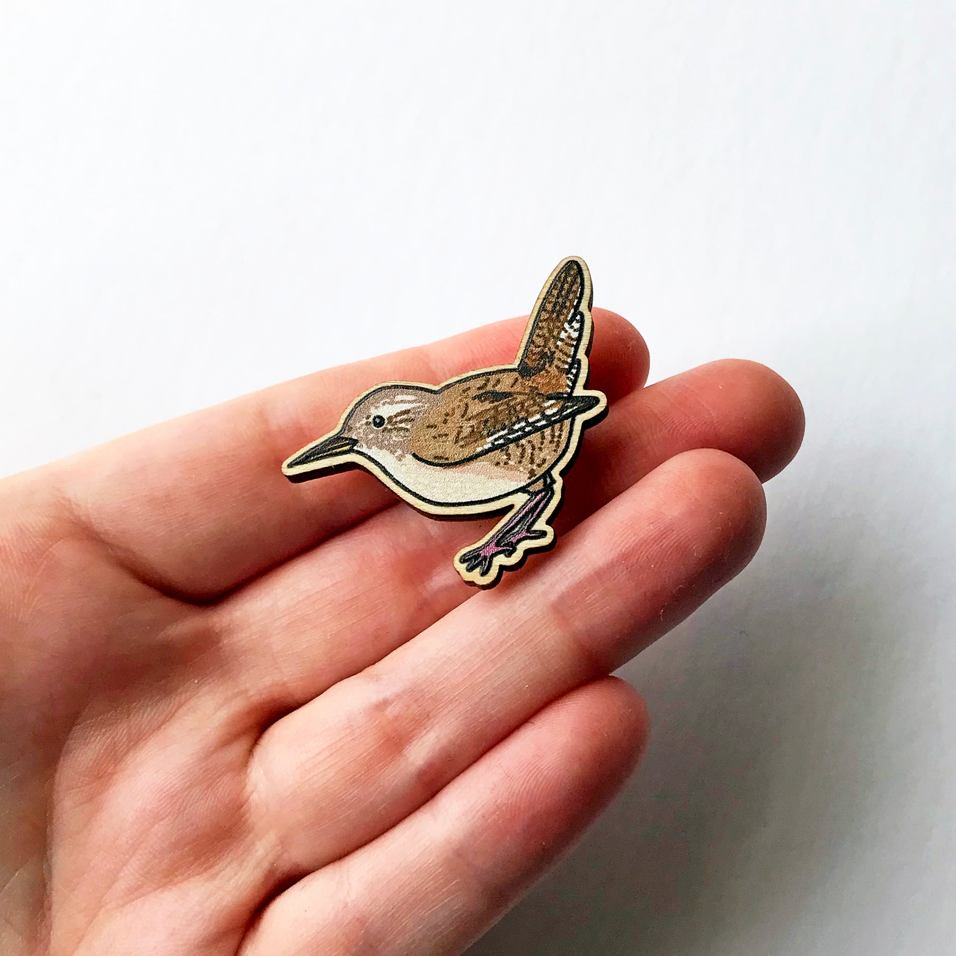 A laser cut wooden brooch showing an illustration by Loadofolbobbins of a wren. The brooch is held in a hand for scale.