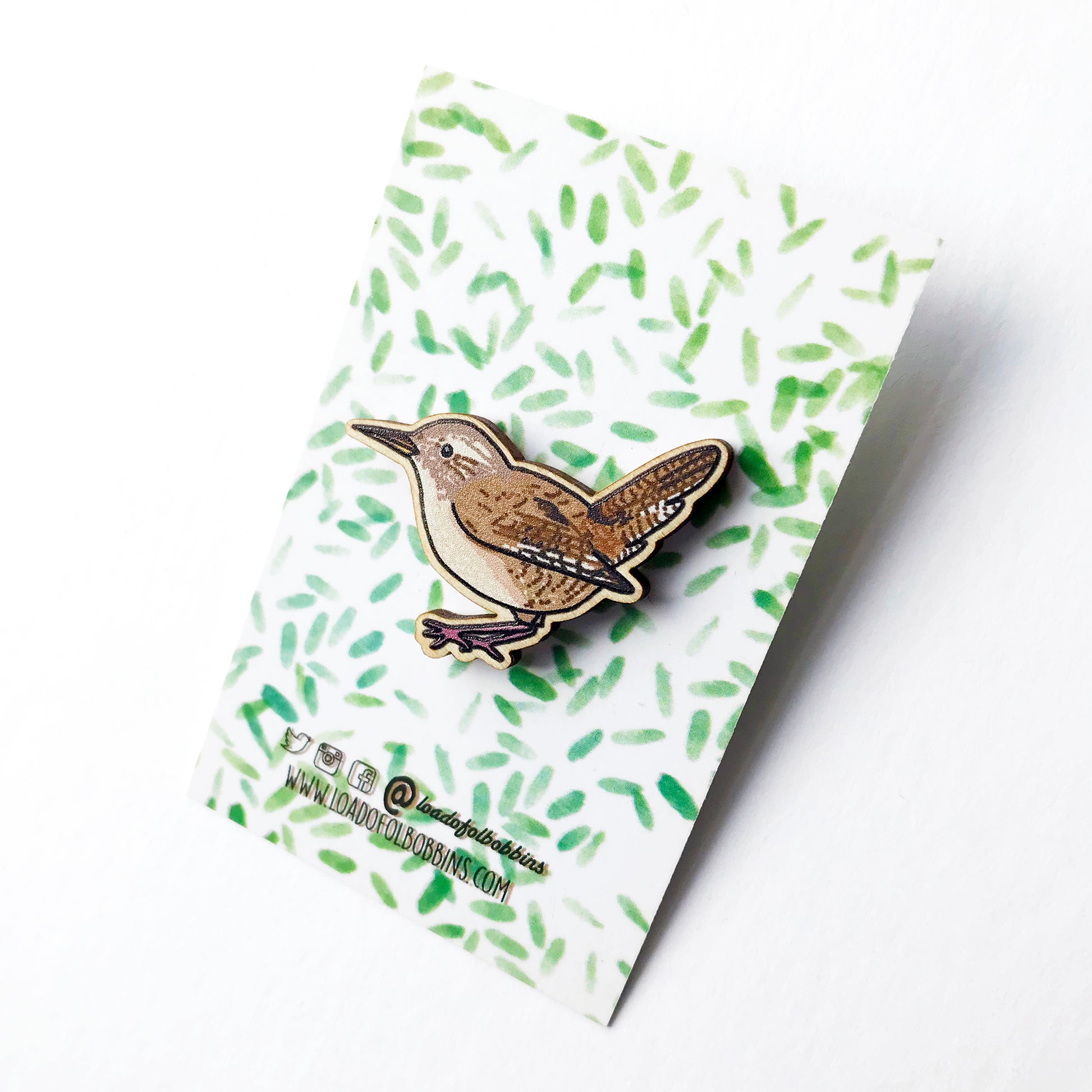 A laser cut wooden brooch showing an illustration by Loadofolbobbins of a wren. The brooch is mounted on a backing card with a repeat abstract watercolour leaf pattern.